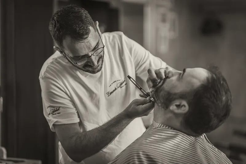 Un barbero sonríe mientras arregla con navaja la barba de un cliente