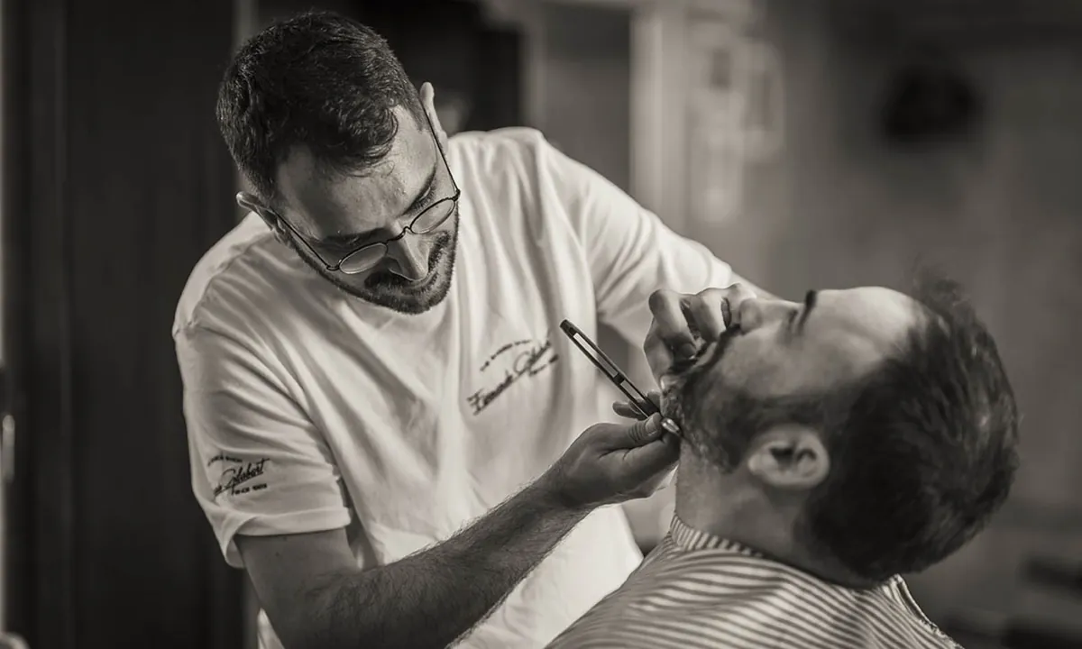 Barbero con gafas arreglando con navaja la barba de un cliente
